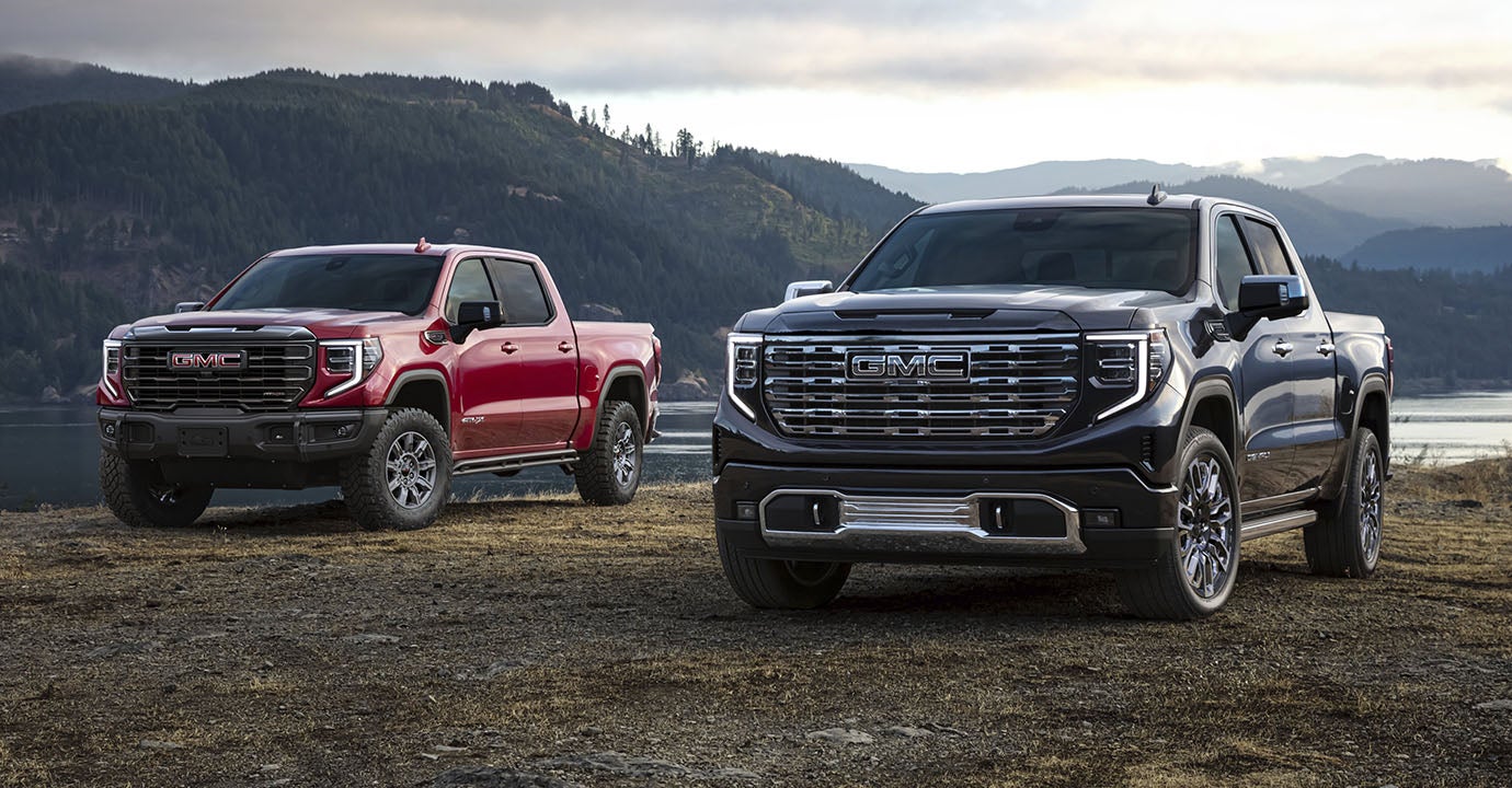 2 high end GMC Sierras, one black, one red. Parked on the bank of a lake in front of some wooded hills.