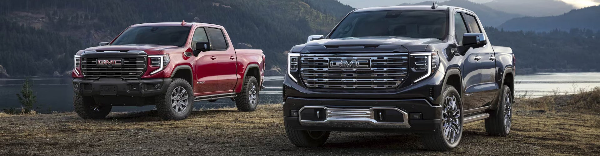 Two GMC pickup trucks, one red and one black, are parked on a scenic lakeside with mountains in the background, conveying strength and adventure.
