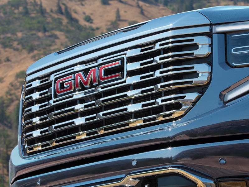 Close-up of a GMC vehicle’s chrome grille with a bold red GMC logo, reflecting sunlight. A rugged, mountainous landscape is visible in the background.