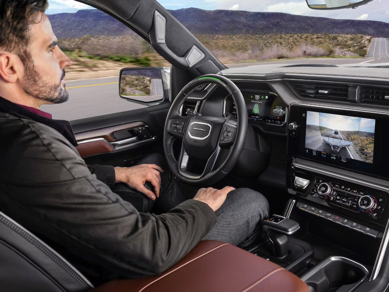 Man seated in a self-driving car interior with hands off the steering wheel, digital dashboard, and touchscreen showing rear camera view.